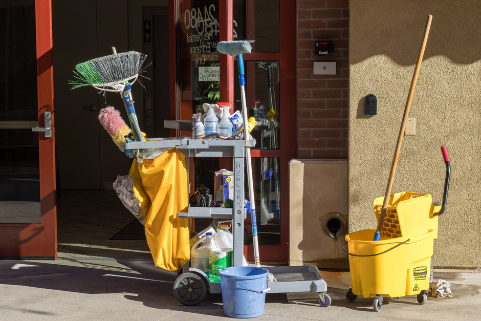 A cleaning cart and equipment outside the entrance of a commercial office building, with a yellow mop bucket that contains a mop with a pink handle, a yellow floor scrubber, and a grey cleaning trolle